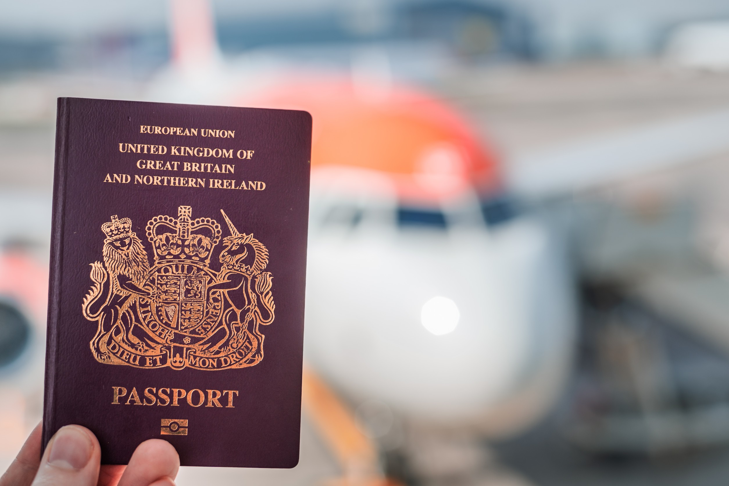 A Red British Passport Held Up Against A Background Of A Generic Plane On A Bright Sunny